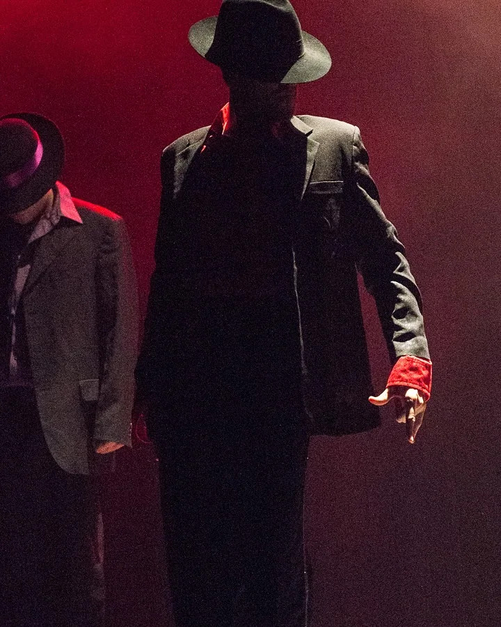 Bailarín de Jackson Project con sombrero, traje negro y puño rojo bajo luces rojas