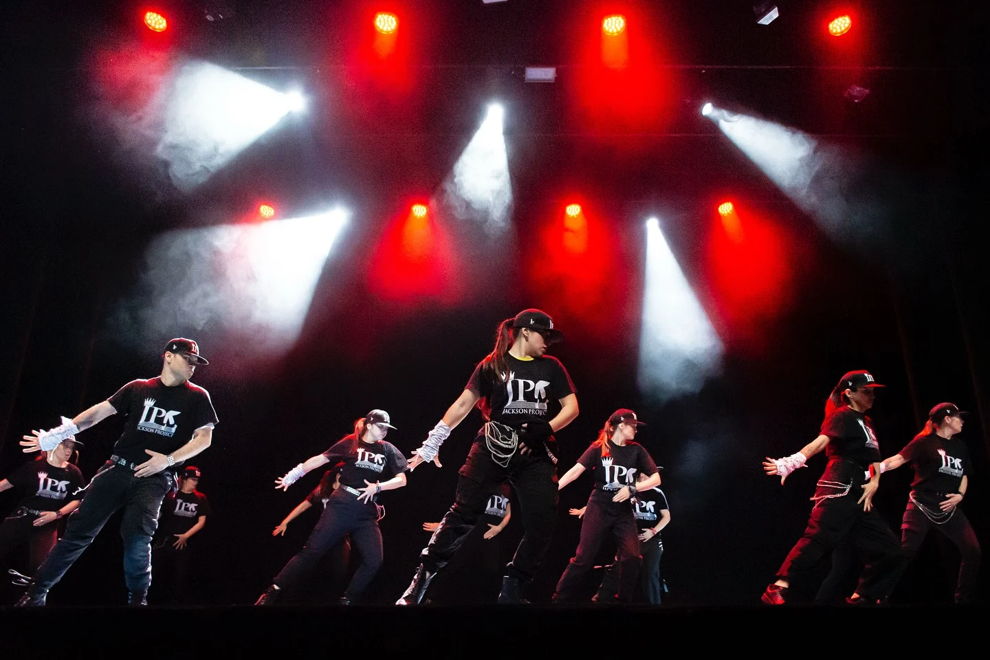 El elenco baila en formación bajo haces de luz roja y blanca