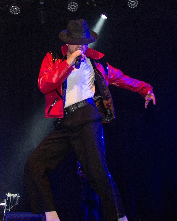 Intérprete con chaqueta roja y sombrero negro en una performance bajo luces azules y blancas